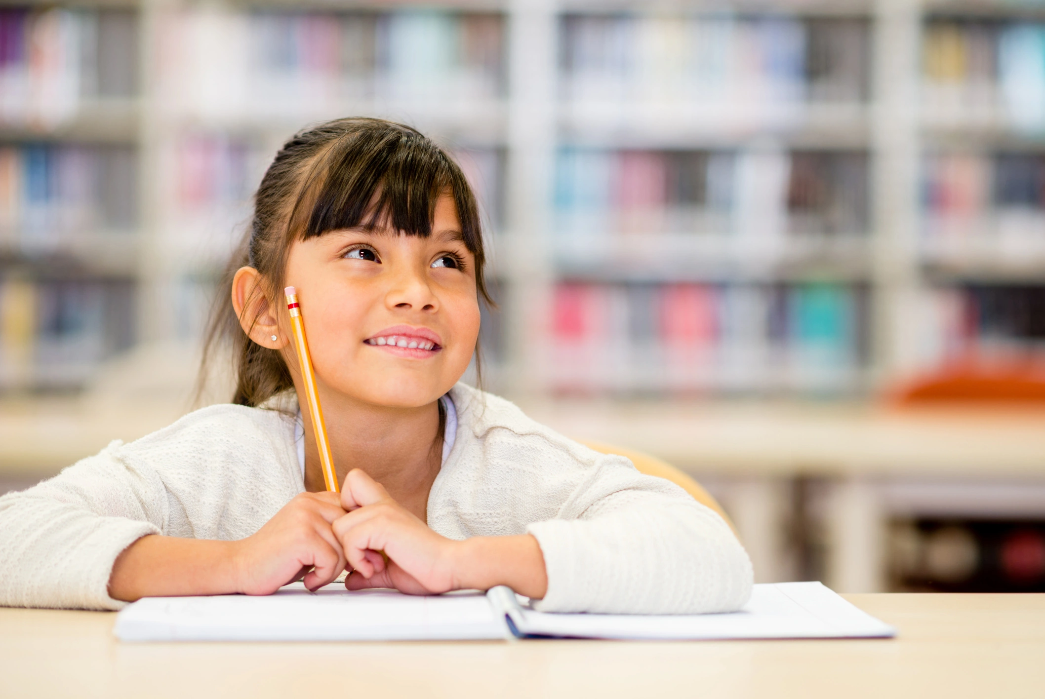 A young girl sits at a table in a library, smiling and looking up while holding a pencil, with an open notebook in front of her—ready to explore new ideas with Online Learning Solutions for K-12. Blurred shelves of books form the background.