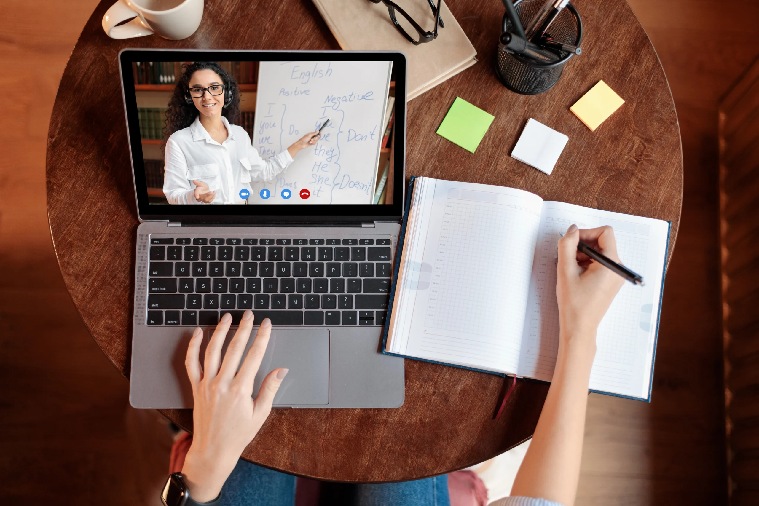 A person takes notes in a notebook while watching an online lesson with a teacher on a laptop. Showcasing Online Learning Solutions for K-12, the scene is set on a round wooden table with stationery and a whiteboard of English words.